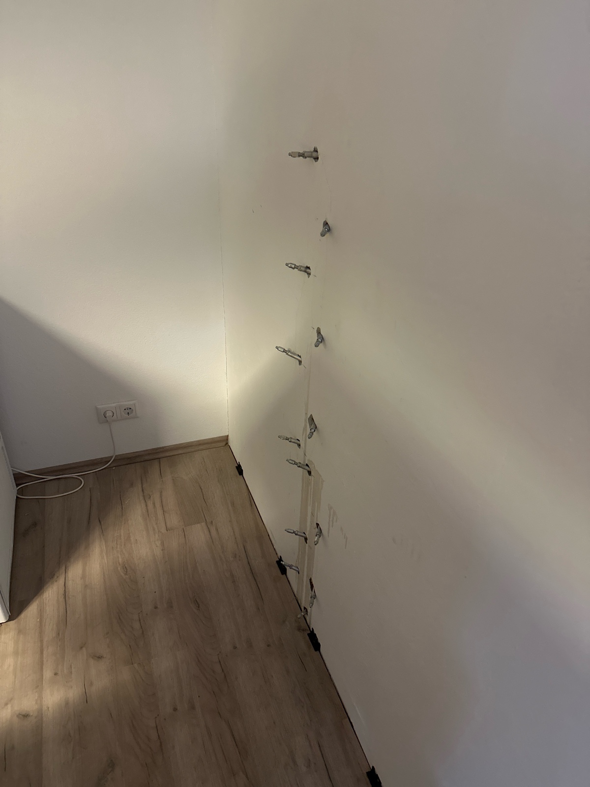 Weiß-beige Wand mit vertikalem, harzverpresstem Riss; mehrere Bohrpacker stecken in der Fuge, am Boden schwarze Abdeckstopfen, darüber ein heller Laminatfußboden.
