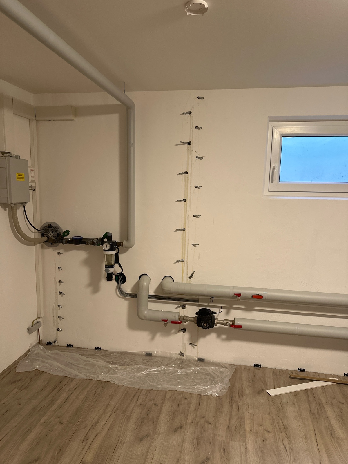 Technikraum im Keller: hell gestrichene Wand mit senkrechter und bodennaher Reihe eingedrehter Bohrpacker, davor verlaufen graue Rohrleitungen mit Absperrarmaturen; auf dem Laminatboden liegt eine schützende Folie.