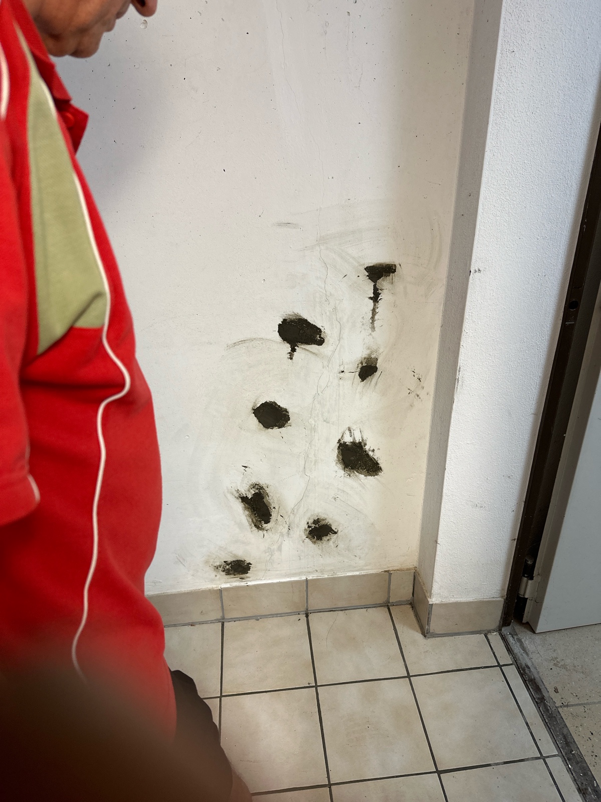 Weiß verputzte Kellerwand neben einer geöffneten Tür; sechs rund ausgespachtelte Injektionsstellen verlaufen senkrecht über der Sockelleiste, davor ein beiger Fliesenboden und am linken Bildrand der rote Arbeitsanzug eines Monteurs.