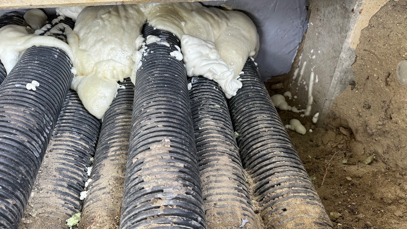 Mehrere parallel liegende, schwarze Leerrohre mit Rippenprofil, teilweise von bräunlichem Erdreich umgeben; aus der Wandöffnung quillt voluminöser, hellbeiger PU-Schaum über die Rohrstränge und dichtet sie ab.