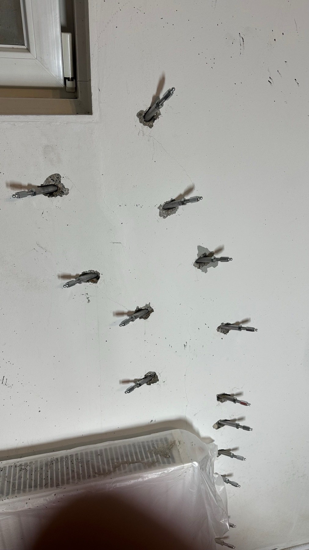 Weiße Innenwand unter einem kleinen Kellerfenster; in zwei versetzten Spuren stecken rund ein Dutzend graue Injektionspacker entlang eines vertikalen Risses, unterhalb ein abgeplanter Heizkörper, Boden mit Schutzfolie abgedeckt.