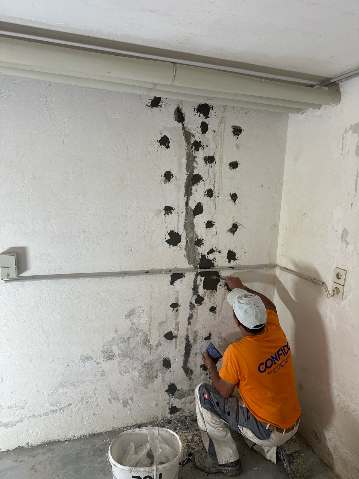 Kellerwand während der Sanierung: Ein Handwerker im orangefarbenen „CONCRET“-Shirt kniet vor zwei senkrechten Risslinien, an denen dicht gesetzte Bohrpacker mit dunklem Harz- bzw. Mörtelauftrag zu sehen sind; weiß gestrichene Wand, horizontale Rohrleitung und Eimer mit Spachtelmasse auf dem gefliesten Boden.