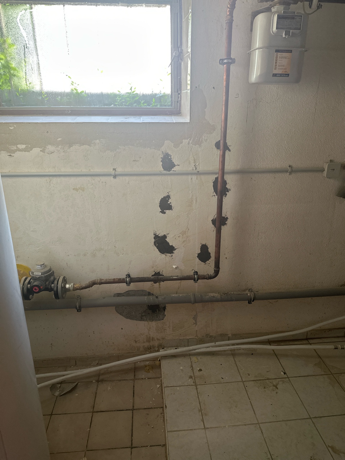 Derselbe Wandabschnitt nach der Injektion: unter einem kleinen Kellerfenster zieht sich eine vertikale Spur grob verspachtelter Bohrlöcher vom Boden bis zur Decke; beige-graue Wand, quer verlaufende Rohre und leicht verschmutzter Fliesenboden im Vordergrund.
