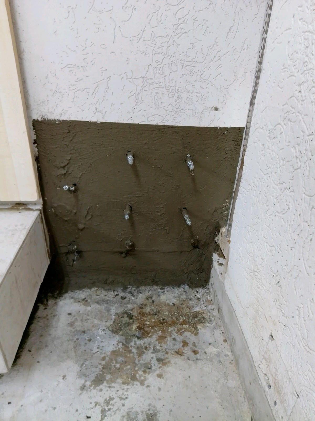 Ecke eines Außenwandfußes: unterer Bereich frisch mit braun-grauer Dichtungsschicht überzogen, mehrere eingedrehte Injektionspacker sitzen rasterförmig darin; weiß verputzte Wand darüber, Betonboden mit feuchten Verfärbungen im Vordergrund.