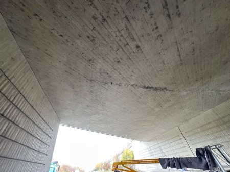 Unteransicht einer Straßenbrücke aus Sichtbeton; in der Mitte zieht sich ein dunkles, längs verlaufendes Rissband über die Deckplatte, am unteren Bildrand sind ein gelber Baggerarm und eingerüstete Pfeiler der Baustelle zu sehen.