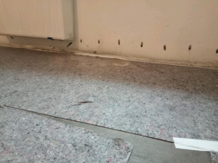 Weiß verputzte Wand mit horizontaler Linie frisch verschlossener Injektionsbohrungen; entlang der Boden-Wand-Fuge eine helle Dichtstoffraupe, auf dem grauen Schutzvliesboden liegt ein abgeschnittener Kunststoffstreifen.