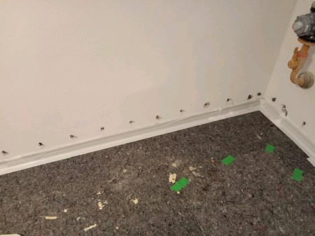 Kellerraumecke mit weiß gestrichener Wand und Sockelleiste; eine Reihe offener Bohrlöcher verläuft knapp über dem Boden, graues Schutzvlies mit grünen Markierungsstreifen sowie Holz-/Putzsplittern liegt auf dem Boden, oben rechts ragt ein Wasser- bzw. Heizungsventil ins Bild.