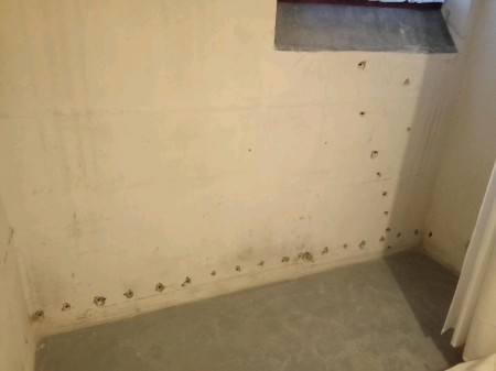 Ecke einer unbehandelten Betonwand im Keller; eine horizontale und eine vertikale Reihe kleiner Bohrlöcher markieren den Wandfuß und die Seitenwand, der blanke Estrichboden ist sichtbar.