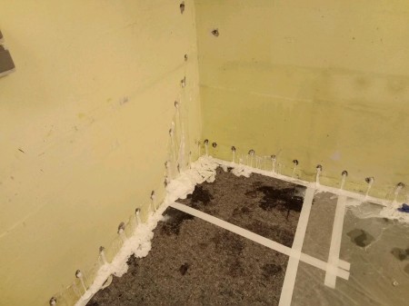 Kellerecke mit gelb gestrichener Wand; entlang des Wandfußes mehrere schlanke Injektionsdübel und aufgesprühter weißer Dichtungsschaum, darunter Folie mit gekreuztem Klebeband und dunklem Feuchtigkeitsfleck auf dem Boden.