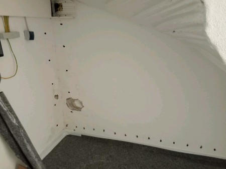Weiß gestrichene Kellerecke unter einer Treppenwange; quer und senkrecht verlaufende Reihe kleiner Bohrlöcher nahe dem Boden, größere aufgestemmte Stelle im Putz sowie sichtbare Kabel an der Wand, grauer Teppichboden im Vordergrund.