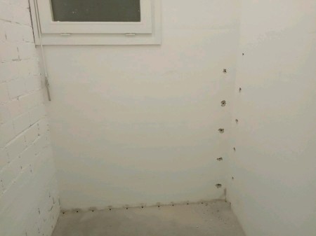 Weiß gestrichene Raumecke unter einem kleinen Fenster; entlang der Boden-Wand-Fuge und senkrecht an der rechten Wand sind mehrere Bohrlöcher gesetzt, roher Betonboden vorne sichtbar.
