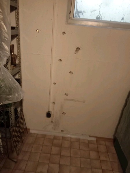 Kellerraum mit beigefarbenem Fliesenboden; an der weiß gestrichenen Wand unter einem kleinen Fenster verläuft eine senkrechte Reihe Bohrlöcher neben einem schmalen Kabelkanal, mehrere größere Ausbrüche im Putz sind sichtbar.