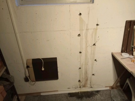 Weiße Betonwand unter Kellerfenster mit senkrecht angeordneten, ausgebrochenen Bohrlöchern; bräunliche Wasserlaufspuren ziehen sich von den Löchern bis zum feuchten Estrichboden hinab, links unten hängt eine schwarze Anschlussbox.