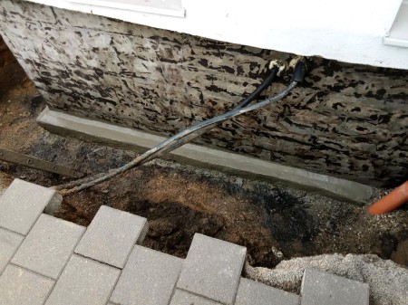 Freigelegte Kelleraußenwand mit beschädigter Bitumenbeschichtung; neue Beton-Hohlkehle am Sockel, Leitungsrohr tritt aus der Wand, davor ausgehobenes Erdreich und lose Pflastersteine.