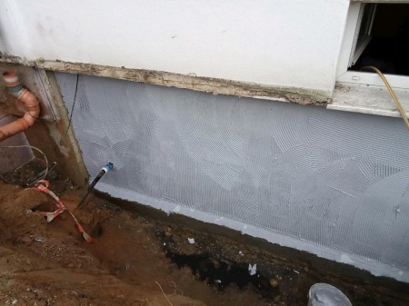 Sanierte Kelleraußenwand: hellgraue Dichtschlämme mit Armierungsgewebe und abgedichteter Rohrdurchführung, frische Hohlkehle am Wandfuß, Boden noch offenliegend.